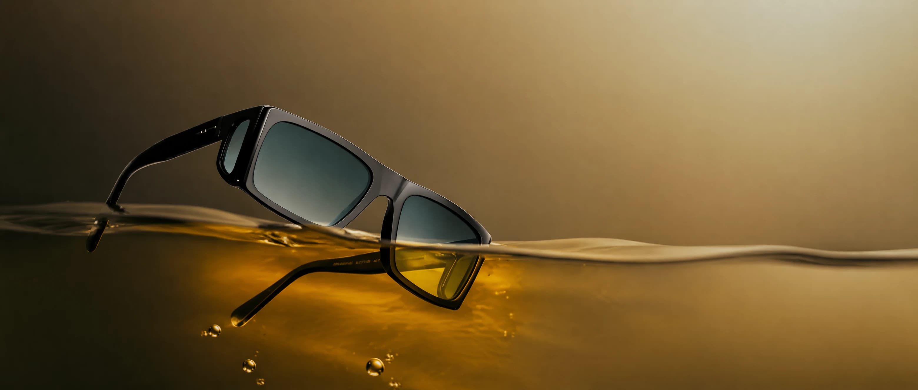 Eyewear hero — black sunglasses half-submerged in golden liquid, editorial product shot